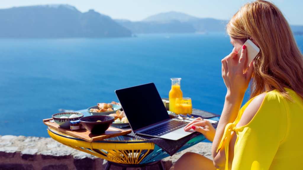 Woman on vacation eating seaside breakfast while taking a call and working on her laptop.