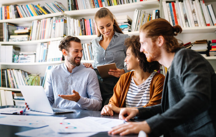 Four professionals collaborating around a table with laptops and documents in a well-lit, book-lined office space