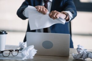 Businessman tearing piece of paper with laptop, glasses, crumpled papers, and coffee on desk